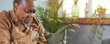 An older man sitting on his couch, clutching his chest in pain.