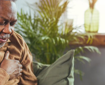 An older man sitting on his couch, clutching his chest in pain.