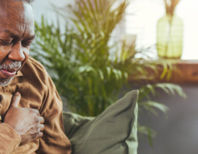 An older man sitting on his couch, clutching his chest in pain.