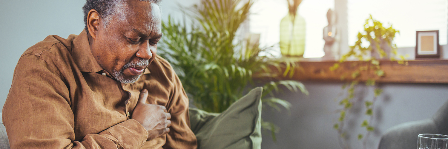 An older man sitting on his couch, clutching his chest in pain.