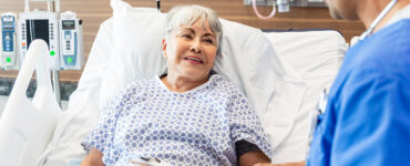 An older woman laying in a hospital bed, speaking to her doctor while smiling.