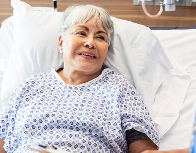 An older woman laying in a hospital bed, speaking to her doctor while smiling.