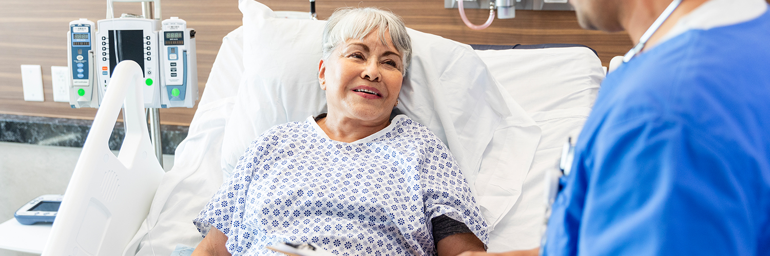 An older woman laying in a hospital bed, speaking to her doctor while smiling.