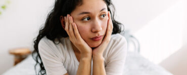 A woman sitting on her bed with her hands around her face, looking stressed.
