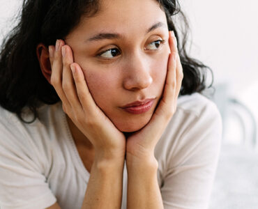 A woman sitting on her bed with her hands around her face, looking stressed.