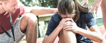 A female athlete who is on the floor, grabbing her leg in pain, with her coach helping her.