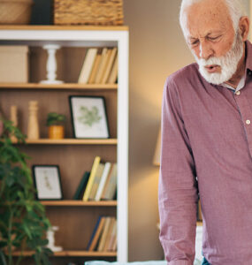 An older man standing in his living room, clutching his hip in pain while leaning on his cane.