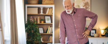An older man standing in his living room, clutching his hip in pain while leaning on his cane.