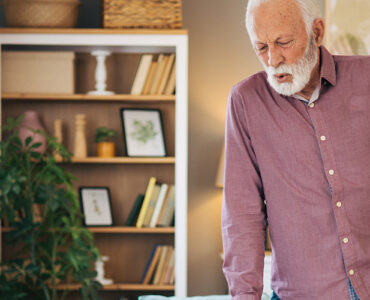 An older man standing in his living room, clutching his hip in pain while leaning on his cane.
