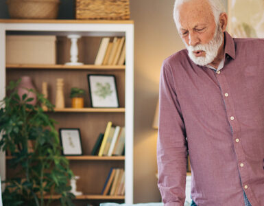 An older man standing in his living room, clutching his hip in pain while leaning on his cane.