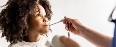 A young girl at a doctor's office, having her temperature taken while looking at the physician's face.