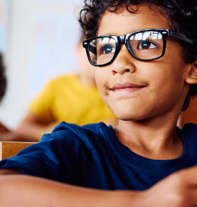 A young boy with glasses sitting in his classroom, looking at something in the distance with a slight smile on his face.