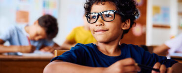 A young boy with glasses sitting in his classroom, looking at something in the distance with a slight smile on his face.