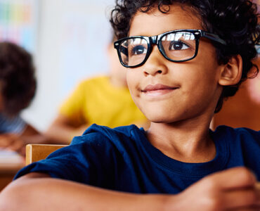 A young boy with glasses sitting in his classroom, looking at something in the distance with a slight smile on his face.
