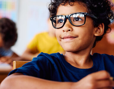 A young boy with glasses sitting in his classroom, looking at something in the distance with a slight smile on his face.