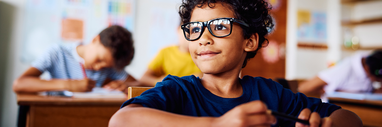 A young boy with glasses sitting in his classroom, looking at something in the distance with a slight smile on his face.