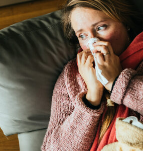 A woman lying on our coach with a blanket over her, while blowing her nose.