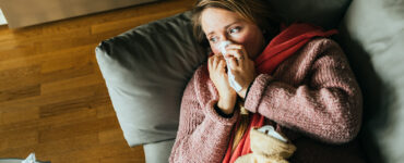 A woman lying on our coach with a blanket over her, while blowing her nose.