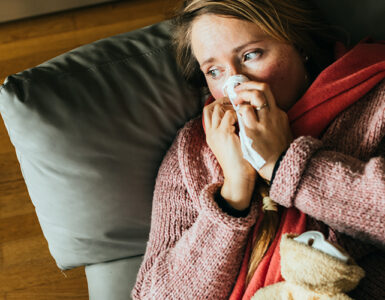A woman lying on our coach with a blanket over her, while blowing her nose.
