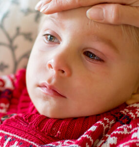 A young boy looking sick, with red eyes and a frown on his face, while his mother feels his forehead.