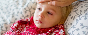 A young boy looking sick, with red eyes and a frown on his face, while his mother feels his forehead.