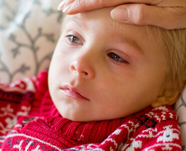 A young boy looking sick, with red eyes and a frown on his face, while his mother feels his forehead.