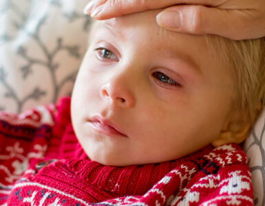 A young boy looking sick, with red eyes and a frown on his face, while his mother feels his forehead.