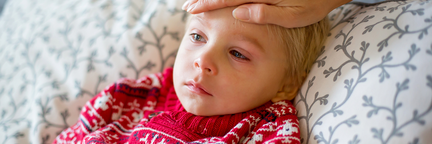 A young boy looking sick, with red eyes and a frown on his face, while his mother feels his forehead.