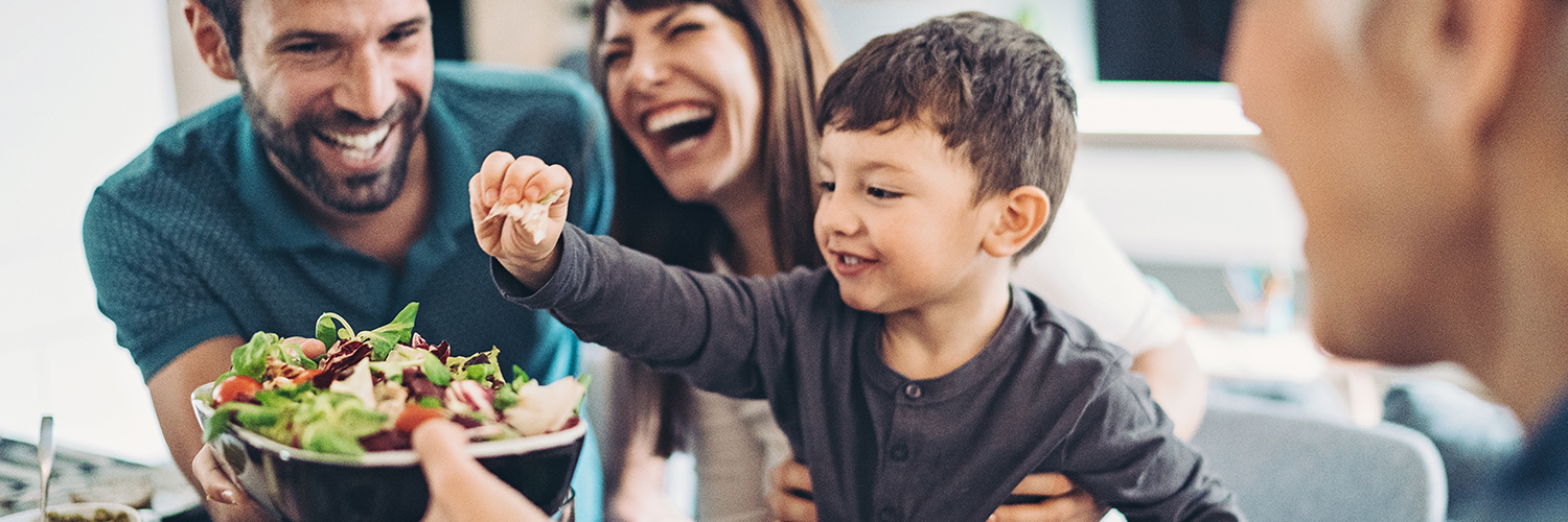 A family working together to prepare a salad, with a little boy helping and smiling.