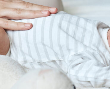 An infant laying down and crying, with mom's hand on his belly.