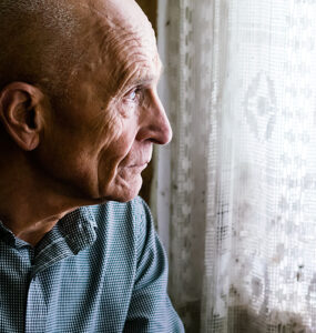 An older man sitting in a chair and looking out a window, looking sad.