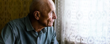 An older man sitting in a chair and looking out a window, looking sad.