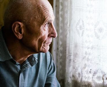 An older man sitting in a chair and looking out a window, looking sad.