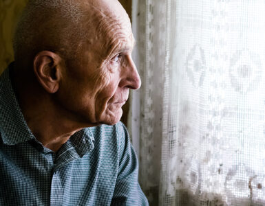 An older man sitting in a chair and looking out a window, looking sad.