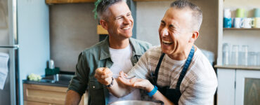 Two middle-aged men cooking dinner together in a kitchen while laughing.