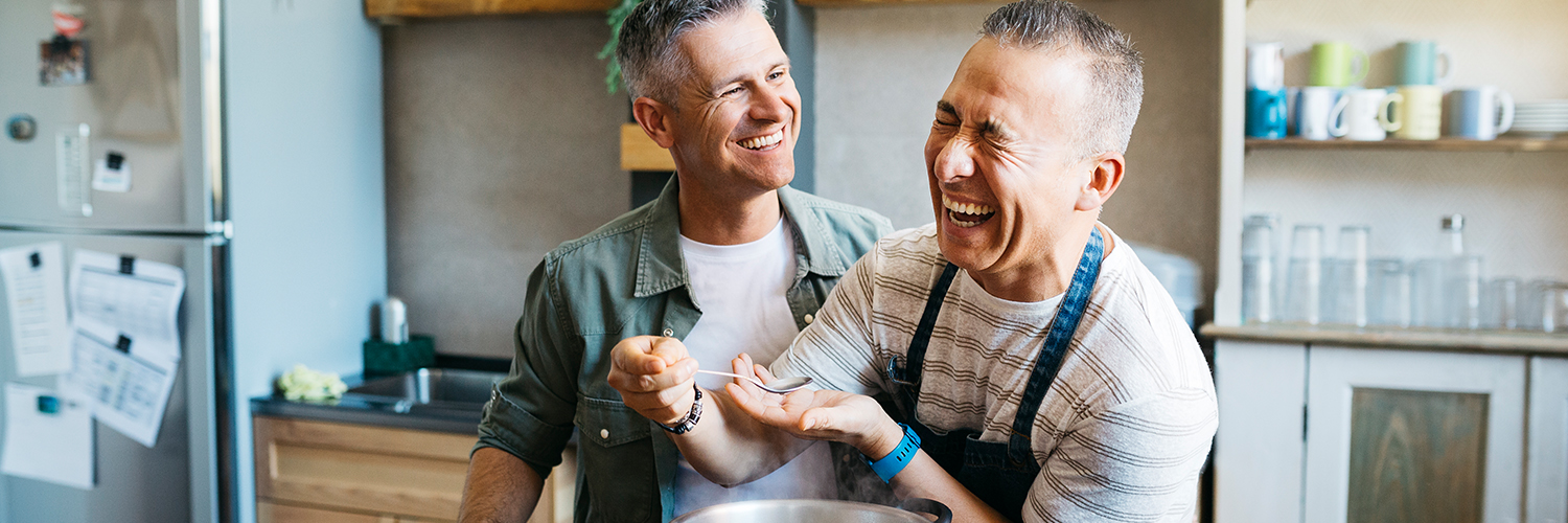 Two middle-aged men cooking dinner together in a kitchen while laughing.