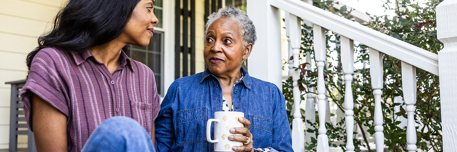 An older black woman sitting next to her daughter on her front stoop, drinking a cup of coffee.