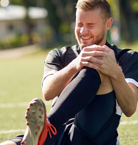 A young man sitting on a soccer field, holding his knee in pain.