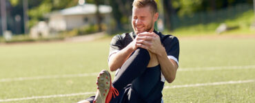 A young man sitting on a soccer field, holding his knee in pain.