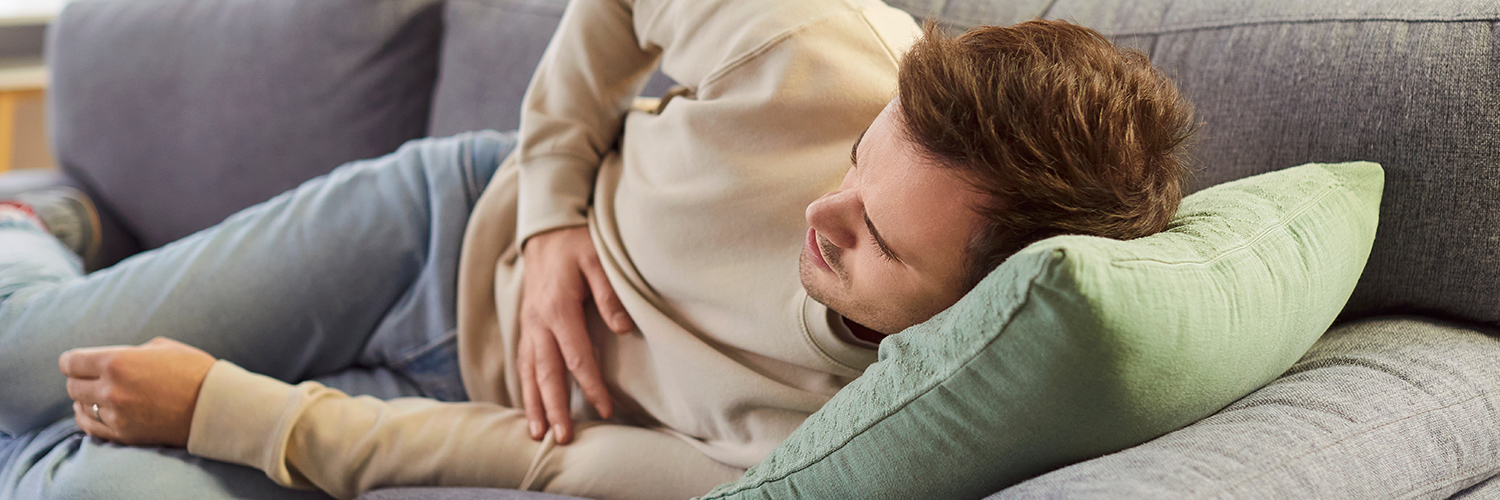 A young man laying on the couch, holding his stomach in pain.