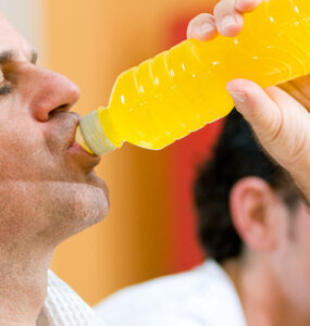 A middle-aged man drinking an energy drink from a bottle while standing in the gym.