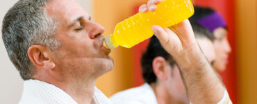 A middle-aged man drinking an energy drink from a bottle while standing in the gym.