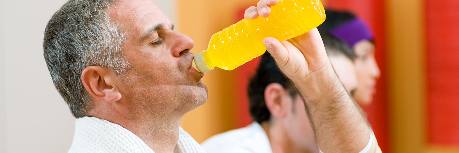 A middle-aged man drinking an energy drink from a bottle while standing in the gym.