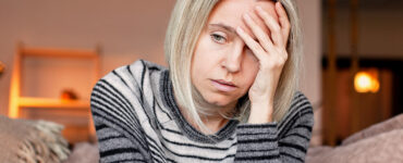 A grieving woman sitting on her couch alone, with her hand to her forehead.