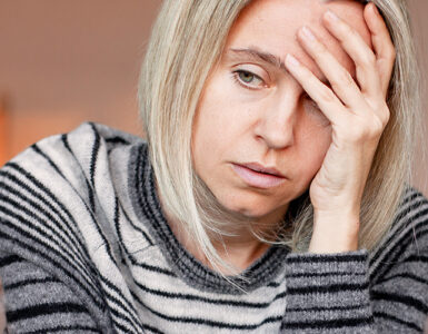 A grieving woman sitting on her couch alone, with her hand to her forehead.