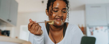 A young black woman leaning over her kitchen island while eating a salad.