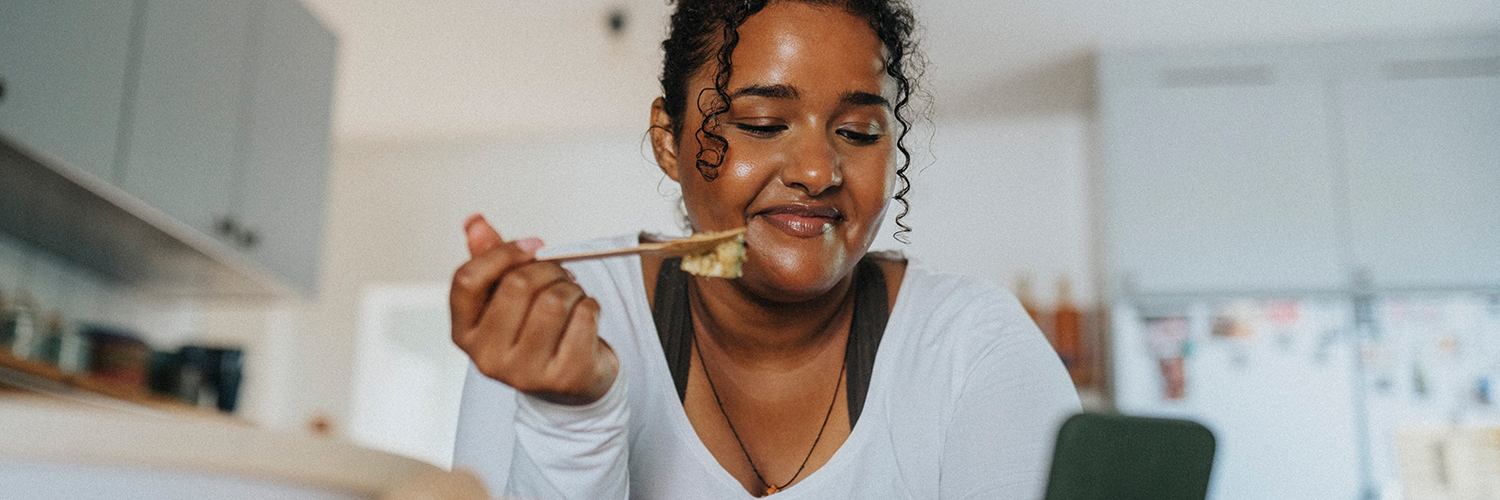 A young black woman leaning over her kitchen island while eating a salad.
