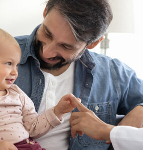 A baby smiling and sitting with her parents, alongside their pediatrician who is touching the baby's hand.