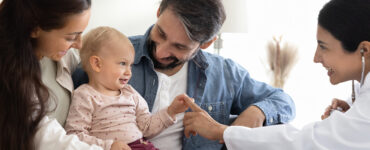 A baby smiling and sitting with her parents, alongside their pediatrician who is touching the baby's hand.
