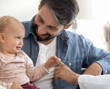A baby smiling and sitting with her parents, alongside their pediatrician who is touching the baby's hand.
